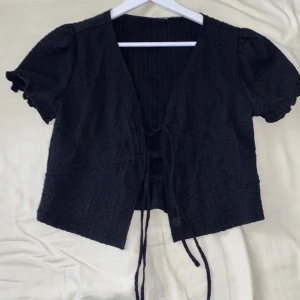 Svart kortärmad bolerotopp med snörning - Supersöt svart bolerotopp med kort puffärm och v-ringning. Toppen har en snygg snörning framtill och är gjord i ett strukturerat, stretchigt tyg. Perfekt att styla över linne eller klänning för en trendig look.