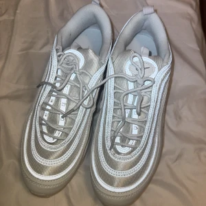 Nike Air Max 97 NYA - Nike Air Max 97 sneakers i en clean silver och vit färg. Skorna har den ikoniska vågiga designen, synlig Air Max-sula och reflekterande detaljer. Ovandelen är i syntet och mesh för en sportig vibe. Perfekta för dig som vill sticka ut med stilrena och futuristiska sneakers. Strlk 44