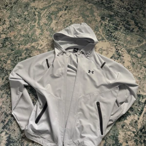 Ljusgrå vindjacka från Under Armour - Snygg ljusgrå vindjacka från Under Armour med huva och svarta dragkedjor på fickorna. Jackan har en sportig look, justerbar dragsko vid huvan och diskret logga på bröstet. Perfekt för blåsiga dagar och outdoor-aktiviteter.