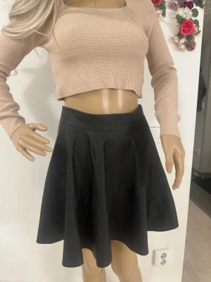 Svart skinnkjol med dragkedja - Säljer en svart, kort skinnkjol med snyggt fall och markerad midja. Kjolen har en guldig dragkedja i sidan som ger en cool detalj. Perfekt för dig som gillar en stilren look med lite edge. Passformen är normal och materialet känns mjukt och följsamt.