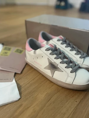 Golden Goose sneakers vit/rosa - Golden Goose Superstar sneakers i vitt skinn med rosa detaljer och grå snörning. Sidan har en ikonisk stjärna i silver och texten GGDB/SSTAR. Insidan är mjuk och sulan är platt med gummimönster. Perfekt för dig som gillar coola och trendiga sneakers.