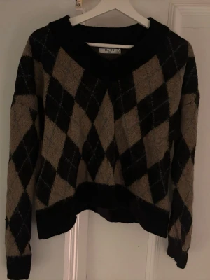 Beige och svart rutig stickad tröja NA-KD - Säljer en mysig stickad tröja från NA-KD med beige och svart argylemönster. Tröjan har v-ringning, långa ärmar och ribbade muddar. Perfekt för lager på lager under hösten, vintern och våren! 