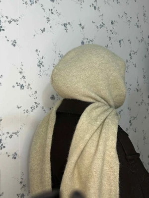 Beige stickad väst med huva(Balaklava) - Säljer en beige stickad väst med huva. Västen är i mjukt och fluffigt material och har en enkel, rak passform utan knappar eller dragkedja. Perfekt att slänga över en tröja för extra värme. Stilren och enkel design som passar till många outfits.