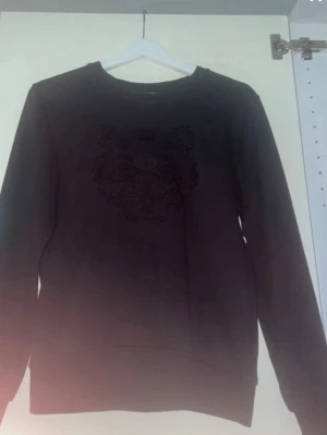 Svart Kenzo sweatshirt med tiger - Säljer en svart sweatshirt från Kenzo i storlek M. Tröjan har det ikoniska tigerbroderiet framtill i svart ton-i-ton och klassisk rund hals. Tillverkad i mjuk bomull och har långa ärmar. Perfekt för dig som gillar streetwear och stilrena detaljer.