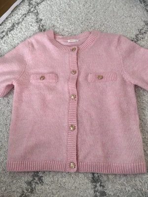 Rosa stickad kofta med guldfärgade knappar - Säljer en ljusrosa stickad kofta från Gina Tricot med rund hals och snygga guldfärgade knappar framtill. Koftan har två dekorativa fickor på bröstet och ribbade kanter vid ärmslut och nederkant. Perfekt för att addera lite färg till din outfit!