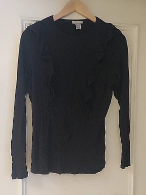 Svart långärmad topp med volang - Säljer en svart långärmad topp från H&M med volangdetaljer längs framsidan och axlarna. Toppen har rund hals och är gjord i mjukt, stretchigt material som sitter bekvämt. Perfekt för dig som gillar stilrena plagg med en twist.