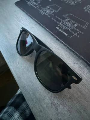 Svart Ray-Ban solglasögon - Ett par svarta Ray-Ban solglasögon med klassisk Wayfarer-form. Glasen är mörka och bågarna är mattsvarta med Ray-Ban-logga på sidan och i glaset. Perfekt accessoar för att höja din stil och skydda ögonen mot solen.