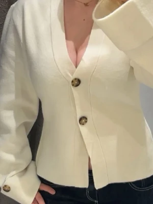 Vit kofta/cardigan från ginatricot - Snygg vit kofta med v-ringning och stora bruna knappar framtill och vid ärmsluten. Koftan har lång ärm, breda muddar och en rak, något croppad passform. Perfekt för lager-på-lager och enkel att styla till olika outfits.