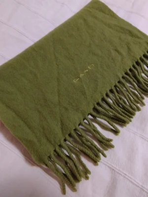 Grön halsduk i lammull från Sand - Säljer en grön halsduk från Sand i fin lammull. Halsduken har klassisk design med tvinnade fransar i kanterna och broderad logga. I fint använt skick.