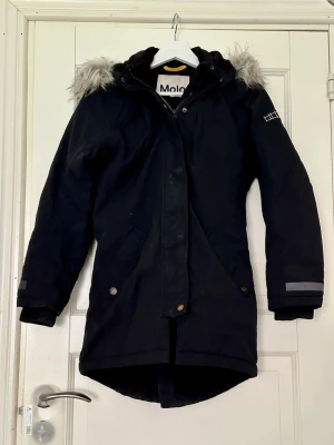 Svart parkajacka med fuskpäls från Molo - Svart parkajacka från Molo i storlek 152. Jackan har huva med grå fuskpäls, dragkedja och knappar framtill samt reflexdetaljer på ärmarna. Tillverkad i återvunnet material och fodrad med 3M Thinsulate för extra värme. Perfekt för kalla dagar.