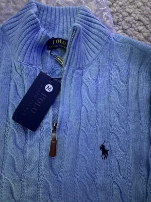 Ljusblå stickad half zip från Polo Ralph Lauren - Säljer en ljusblå stickad tröja från Polo Ralph Lauren med half zip och kabelstickat mönster. Tröjan har ribbad krage, muddar och det klassiska broderade logot på bröstet. Perfekt för dig som gillar stilren och bekväm design.