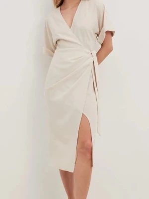 Beige omlottklänning med slits - Snygg beige omlottklänning med v-ringning och knyt i midjan. Klänningen har korta ärmar och en elegant slits framtill som ger en cool touch. Materialet känns mjukt och lätt, perfekt för varmare dagar. Stilren design som är enkel att matcha med accessoarer.