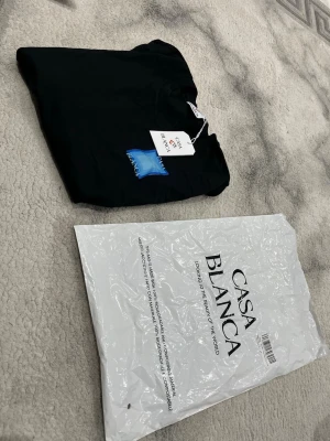 Svart Casa Blanca t-shirt med blå tryck - Svart t-shirt från Casa Blanca med ett coolt blått tryck på bröstet där det står 'CASA BLANCA'. Klassisk rund hals och korta ärmar.l som känns skön mot huden. Perfekt för dig som gillar stilrena plagg med en twist. Priset kan självklart diskuteras 😊. T-shirten är rätt så liten i storlek så rekommenderas till den som har storlek M (medium)