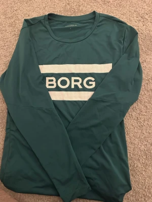 Grön långärmad träningströja Björn Borg - Snygg grön långärmad träningströja från Björn Borg med vit logga på bröstet. Tillverkad i funktionsmaterial HYDRO-PRO som transporterar bort fukt. Perfekt för dig som gillar sportiga och stilrena plagg.