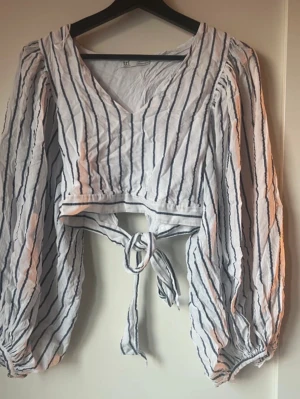 Randig omlottopp med knyt - Säljer en vit och marinblå randig omlottopp från Zara med ballongärmar och knyt i midjan. Toppen har v-ringning och croppad passform, samt långa ärmar med resår vid handlederna. Perfekt för dig som gillar trendiga detaljer och luftig stil.