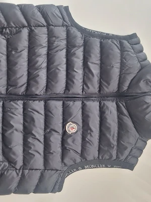 Svart dunväst från Moncler - Snygg svart dunväst från Moncler med quiltad design och dragkedja framtill. Västen har ett klassiskt Moncler-märke på bröstet och diskreta logodetaljer längs ärmöppningarna. Perfekt lager för kyligare dagar.