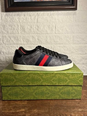 Gucci Ace  - Säljer ett par snygga Gucci sneakers i mörkgrått med klassiskt GG-mönster och breda ränder i rött och marinblått på sidan. Skorna har vit platt sula med Gucci-logga undertill och svarta skosnören. Materialet är canvas med detaljer i skinn.