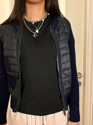 Mörkblå Moncler kofta med quiltad front, stl S - Snygg mörkblå kofta i storlek S med stickade ärmar och rygg samt quiltad front i syntetmaterial. Koftan har tvåvägsdragkedja och praktiska fickor framtill. Perfekt för lager-på-lager under höst och vinter. Stilren och modern look som passar till många outfits.