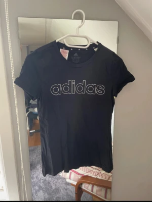 Svart Adidas t-shirt med logga - Svart t-shirt från Adidas med stor vit logga på bröstet och mindre logga i nacken. Klassisk rund hals och korta ärmar. Tillverkad i mjuk bomull som är skön mot huden. Perfekt för dig som gillar sportig och enkel stil.