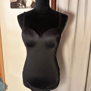 Svart korsettbody med axelband - Säljer en svart korsettbody med justerbara axelband och vadderade kupor. Bodyn har en slät och glansig yta, samt en öppning i ryggen. Perfekt för att skapa en snygg siluett under kläder eller som statement-plagg.