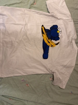 Vit t-shirt KAWS x Andy Warhol Uniqlo - Säljer en vit t-shirt från Uniqlo med KAWS x Andy Warhol-design. Framsidan har en liten gul och blå detalj med Warhols signatur och två kryss, medan baksidan har ett stort tryck med en blå KAWS-figur och en gul banan. Klassisk passform och mjukt bomullsmaterial. Använd två gånger. Kostar 1699 ny