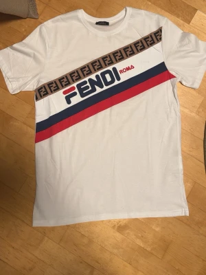 Vit Fendi t-shirt med logga, strl L - Säljer en vit t-shirt från Fendi i storlek L. T-shirten har korta ärmar och är gjord i mjuk bomull. Framsidan har Fendi Roma-logga, röda och blå diagonala ränder samt ett brunt band med Fendi-monogram. Perfekt för dig som gillar streetwear och designer.