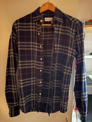 Rutig skjorta John Henric - Blårutig skjorta från John Henric med vita knappar. Skjortan är gjord i mjuk bomull. Mönstret består av breda rutor i blått, grått och tunna röda linjer. Perfekt för dig som gillar stilrena och tidlösa plagg.