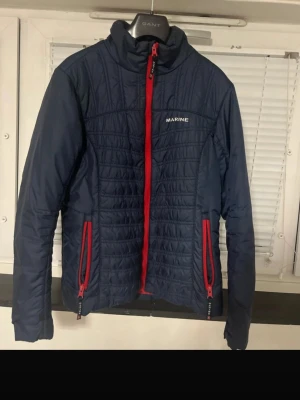 Marine Scandinavian Sportswear Quilted Jacket – Navy Blue & Red - Stylish and lightweight quilted jacket from Marine Scandinavian Sportswear. Classic navy blue color with eye-catching red zippers and details. Perfect as a spring/fall jacket or as a layering piece in colder weather. Comfortable fit, practical zip pockets, and a clean Scandinavian design. Brand: Marine Scandinavian Sportswear Size: XS / EU 34 Color: Navy blue with red details Condition: Very good condition, no stains or damage Style: Casual, sporty, everyday wear Smoke-free and pet-free home. Fe