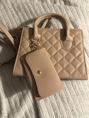 Beige quiltad handväska med plånbok - Säljer en beige handväska med quiltat mönster och guldfärgade detaljer. Väskan har både handtag och axelrem, samt en matchande beige plånbok med guldfärgad knapp. Perfekt för dig som gillar stilrena accessoarer med snygga detaljer. Från new Yorker, aldrig använt. 