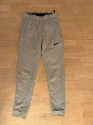 Grå Nike mjukisbyxor Dri-FIT S - Grå mjukisbyxor från Nike med svart logga på benet och elastisk midja med snörning. Tillverkade i Dri-FIT-material som andas och håller dig torr. Byxorna har smal passform, muddar vid bensluten och två sidofickor. Perfekta för chill eller träning.