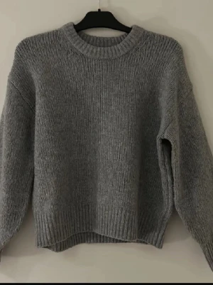 Grå stickad tröja med rund hals - Mysig grå stickad tröja med rund halsringning och ribbade muddar. Tröjan har en loose passform och långa ärmar, perfekt för lager-på-lager under kyliga dagar. Tröjan har några få små rosa fläckar vid halsorngningen kan kanske gå bort vid tvätt men vet ej, skriv för detaljerade bilder