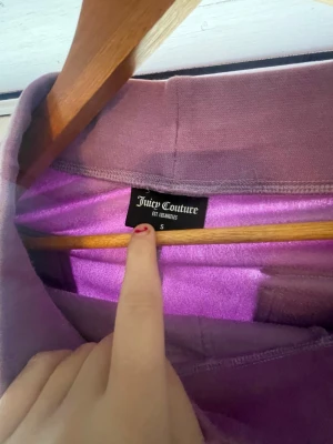 Lila mjukisbyxor Juicy Couture S - Säljer ett par lila mjukisbyxor från Juicy Couture i storlek S. Byxorna har snörning i midjan, stora fickor fram och bak med lock och metallknapp. Materialet är mjukt och skönt, perfekt för chill dagar. Snygg bootcut-modell som ger en avslappnad vibe., en fick knapp är tyvärr borta så säljer denna billigare 