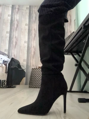 Svarta höga stövletter med klack - Säljer ett par svarta stövletter med spetsig tå och hög smal klack. Skorna är tillverkade i mocka och går upp över knäet. Medföljer extra klacktoppar. Perfekt för dig som vill ha en snygg och stilren look. Oanvända