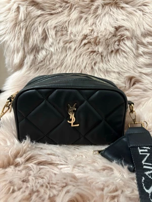Svart axelväska - Säljer en svart quiltad axelväska med guldiga detaljer och logotyp framtill. Väskan har två dragkedjor upptill och en bred axelrem med Saint Laurent-tryck. Materialet är mjukt skinn och väskan har en liten rundad ficka som hänger på sidan.