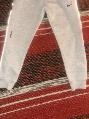 Grå Nike NOCTA mjukisbyxor - Säljer ett par ljusgrå mjukisbyxor från Nike NOCTA med diskret logga på benet och klassisk Nike Swoosh vid höften. Byxorna har resår i midjan och benslut samt reflekterande detaljer längs sidorna. Perfekta för chill eller streetwear.