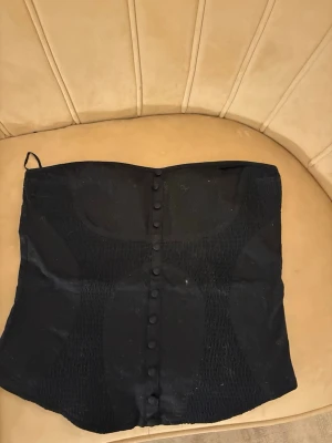 Svart korsettopp/ tubtop från Vila - Säljer en svart korsettopp från Vila i storlek M. Toppen är axelbandslös och har dekorativa knappar framtill samt smockade partier på sidorna för en snygg passform. Perfekt att styla med jeans eller kjol för en trendig look.