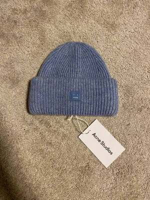 Blå ribbstickad mössa från Acne Studios - Säljer en blå ribbstickad mössa från Acne Studios med klassisk logopatch framtill. Mössan är tillverkad i 100% ull och har en bred uppvikt kant för extra värme. Perfekt för kyliga dagar och snygg att styla till jacka eller hoodie.