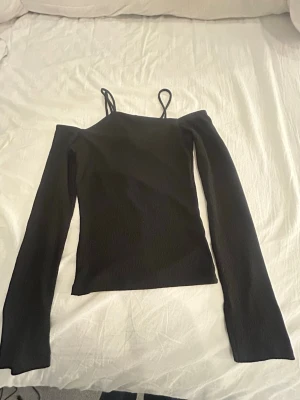 Svart ribbad topp från Gina Tricot - Säljer en svart ribbad topp från Gina Tricot i storlek XS. Toppen har smala axelband och långa ärmar med lätt utsvängda avslut. Materialet är mjukt och stretchigt, vilket gör den bekväm att bära. Perfekt att styla med jeans eller kjol.