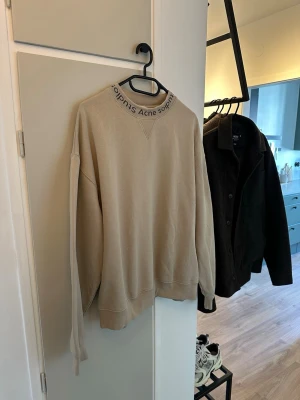 Beige sweatshirt från Acne Studios - Säljer en beige sweatshirt från Acne Studios med tryckt logga runt den höga kragen. Tröjan har lång ärm, loose passform och är tillverkad i mjuk bomull. Perfekt för dig som gillar stilrena och bekväma plagg med en touch av designer.