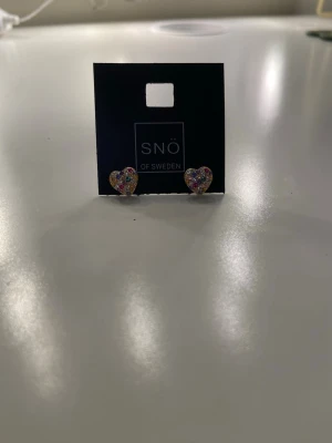 Glittriga hjärtformade örhängen SNÖ - Säljer ett par små hjärtformade örhängen från SNÖ of Sweden. De är guldiga och täckta med glittriga stenar i olika färger som rosa, blå, grön och lila. Perfekt för dig som gillar färgglada accessoarer med lite extra bling.