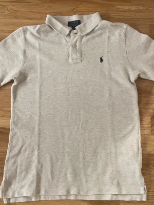 Polo Ralph Lauren Pike - Säljer en klassisk beige pikétröja från Polo Ralph Lauren i limited edition med broderad marinblå logga på bröstet. Tröjan har korta ärmar, krage och två knappar framtill. Utmärkt för finare tillfällen 