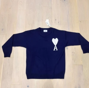 Marinblå stickad tröja från Ami - Snygg marinblå stickad tröja från Ami Alexandre Mattiussi med ett stort vitt hjärta och bokstaven A på bröstet. Tröjan har rund hals, ribbade muddar och lång ärm. Perfekt för dig som gillar stilrena detaljer och vill ha något som sticker ut.