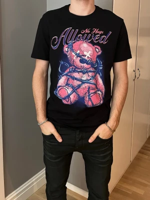 Svart t-shirt med björntryck - Svart t-shirt med färgstarkt tryck av en rosa nallebjörn insnärjd i taggtråd och texten 'No Hugs Allowed' i neonstil. T-shirten har normal passform och korta ärmar. Materialet känns som mjuk bomull och trycket ger en edgy vibe.