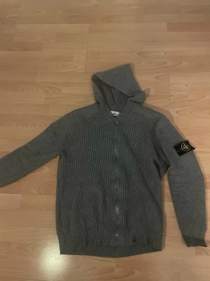 hoodie Stone Island - Säljer en grå ribbstickad hoodie från Stone Island med huva och dragkedja framtill. Tröjan har lång ärm och den klassiska Stone Island-loggan på vänster ärm. Perfekt för dig som gillar streetwear och vill ha något stilrent och bekvämt.