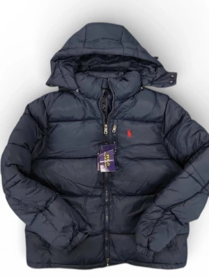 Mörkblå pufferjacka från Polo Ralph Lauren - Snygg mörkblå pufferjacka från Polo Ralph Lauren med huva och dragkedja framtill. Jackan har det klassiska röda Polo-logot broderat på bröstet och är quiltad för extra värme. Perfekt för kalla dagar och har en loose passform.