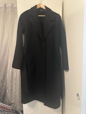 Svart kappa - Svart lång kappa från Vero Moda med klassisk krage och bälte i midjan och två djupa sidofickor. Perfekt för dig som vill ha en stilren och tidlös look under höst och vinter. Kappan är i väldigt bra skick ✨