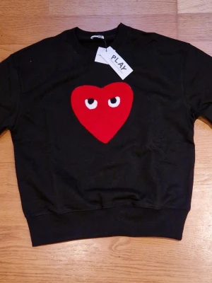 Svart tröja Comme des Garçons Play - Svart tröja från Comme des Garçons Play med ett stort rött hjärta med ögon broderat på bröstet. Rund hals och ribbade muddar. Tröjan har en loose passform och är tillverkad i mjuk bomull. Perfekt statement-plagg för dig som gillar coola detaljer.