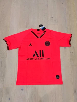 PSG x Jordan röd fotbollströja M - Snygg röd PSG x Jordan fotbollströja i herrstorlek M med svarta detaljer och loggor. Tröjan har v-ringad hals och korta ärmar med svarta ränder vid mudden. Materialet är lätt och andas, perfekt för fotboll eller träning. Officiell produkt med etikett kvar.