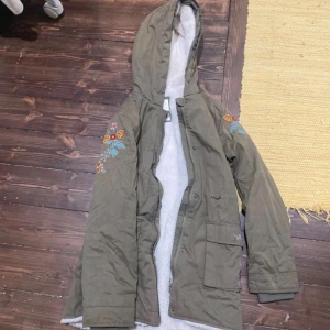 Olivgrön parkas med broderi och luva - Säljer en olivgrön parkas med broderade blommor på ärmarna och mysigt vitt foder. Jackan har luva, dragkedja och stora fickor framtill. Perfekt för dig som vill ha en unik och varm jacka med snygga detaljer.