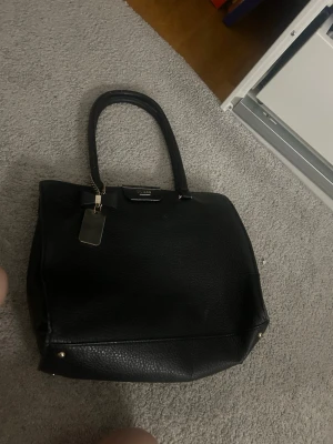 Svart handväska från Guess - Säljer en stilren svart handväsk med grov textur och guldfärgade detaljer. Vet ej om väskan är äkta så darför säljer ja den så billigt. Väskan har flera fack, dragkedjor och en snygg metallbricka med Guess-logga. Perfekt storlek för att få plats med allt du behöver och har både handtag och innerfack.